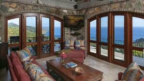 A spacious living room with a large sofa, a coffee table, and a television mounted on a stone wall. The room features large glass doors leading to a balcony with a view of the ocean and hills. - South Shore Villa, SolVillasUSVI (Cruz Bay)