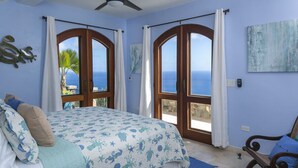 A bedroom with a bed, a ceiling fan, a window with curtains, a view of the sea, and a painting on the wall. - South Shore Villa, SolVillasUSVI (Cruz Bay)