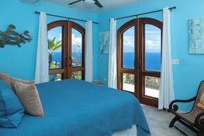A bedroom with a large bed, two windows with curtains, a ceiling fan, and artwork on the walls.