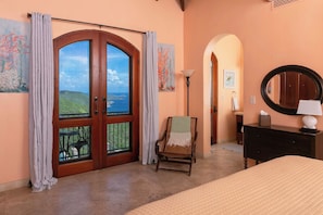 A bedroom with a large window, a bed, a chair, a dresser, and a mirror.