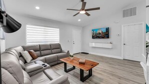 Un salon moderne avec un canapé d’angle, une table basse en bois, un ventilateur de plafond et un tableau représentant un paysage urbain accroché au mur.