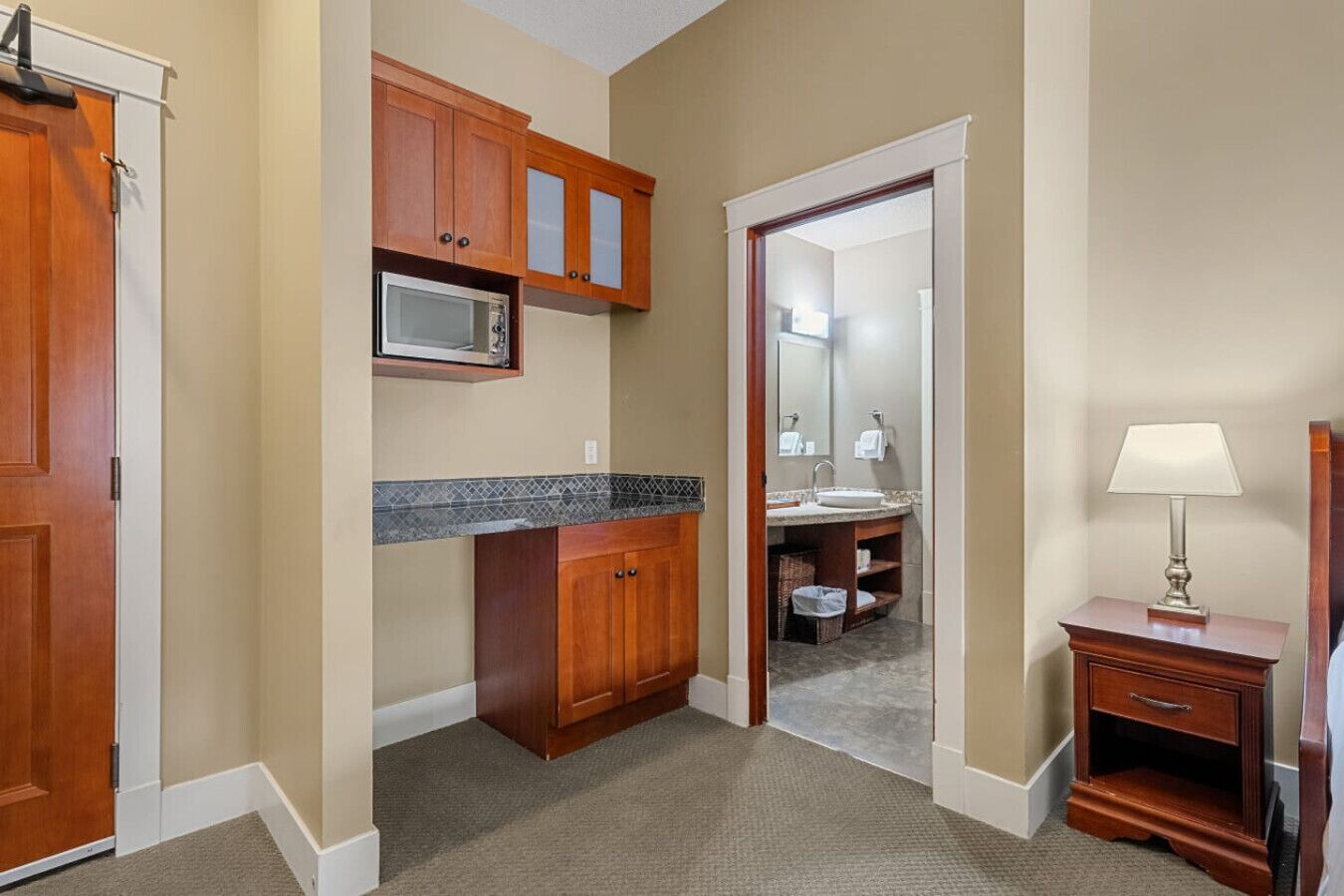 Un baño de hotel con armarios de madera, un microondas, un lavamanos, un espejo y una mesita de noche con lámpara.