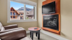 Una sala de estar acogedora con chimenea, un televisor montado arriba y un ventanal que ofrece vistas a una montaña y un edificio de madera.