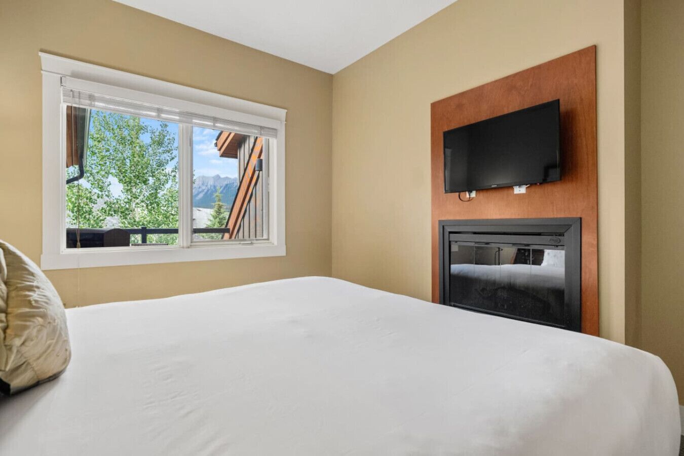 A hotel room with a bed, a TV mounted on a wooden panel, and a window with a mountain view.