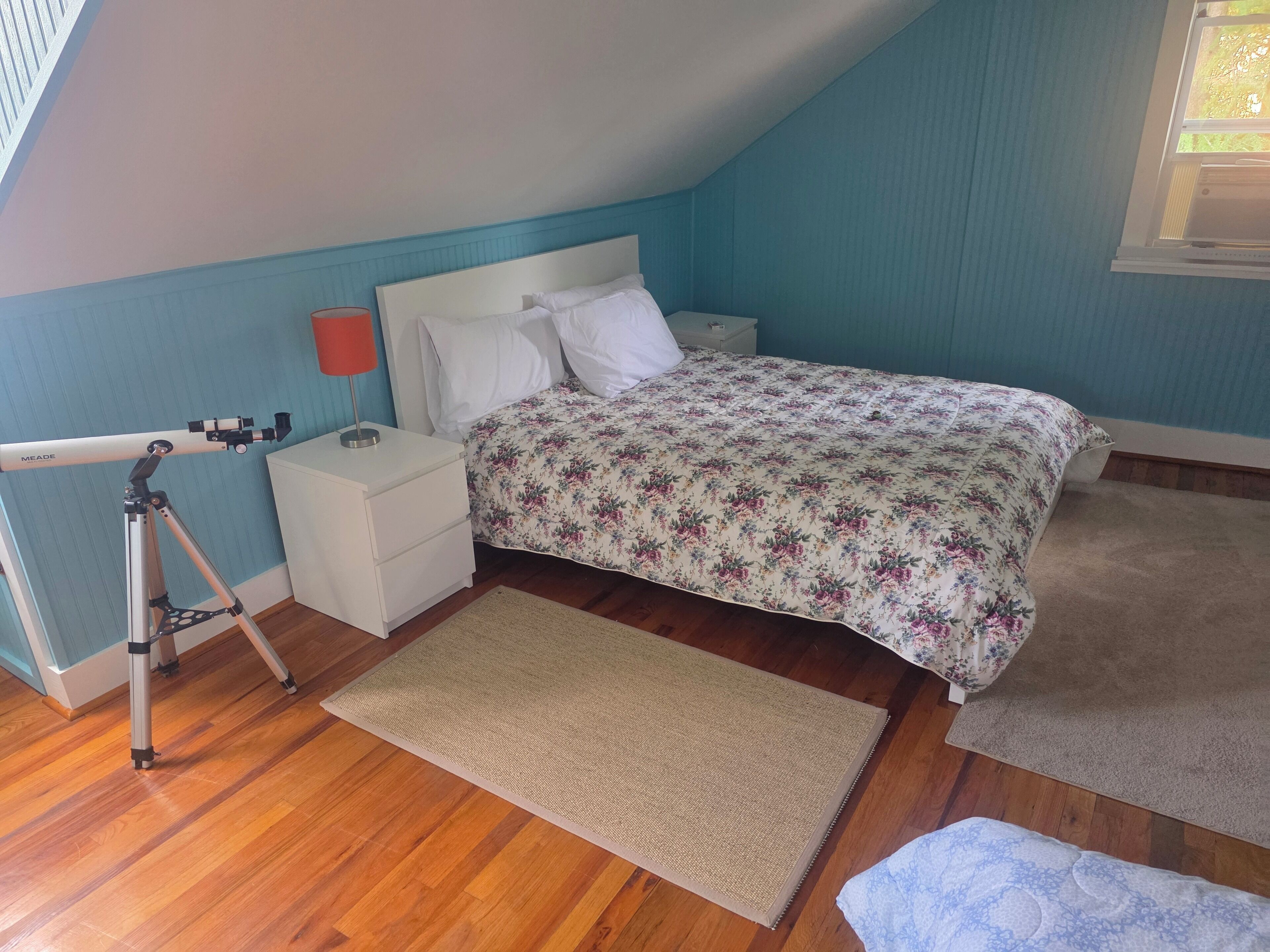 Un dormitorio con cama, mesita de noche, lámpara, telescopio y ventana.