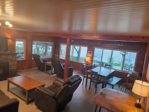 A cozy cabin interior with a stone fireplace, leather recliners, a dining table, and large windows overlooking a lake.
