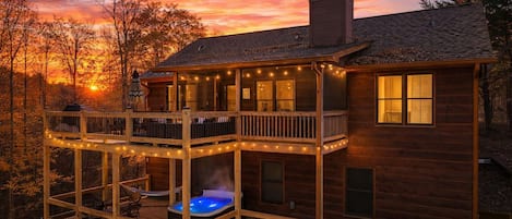 A two-story wooden cabin with a hot tub on the ground floor and string lights on the balcony.
