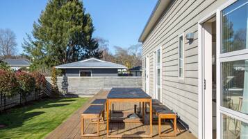 A wooden deck with a table and chairs, a house with a metal roof, and a large tree in the background.