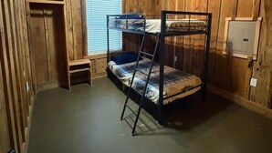Quarto com beliches, paredes de madeira, ventilador de teto, janela com persianas e um pequeno banco de madeira.