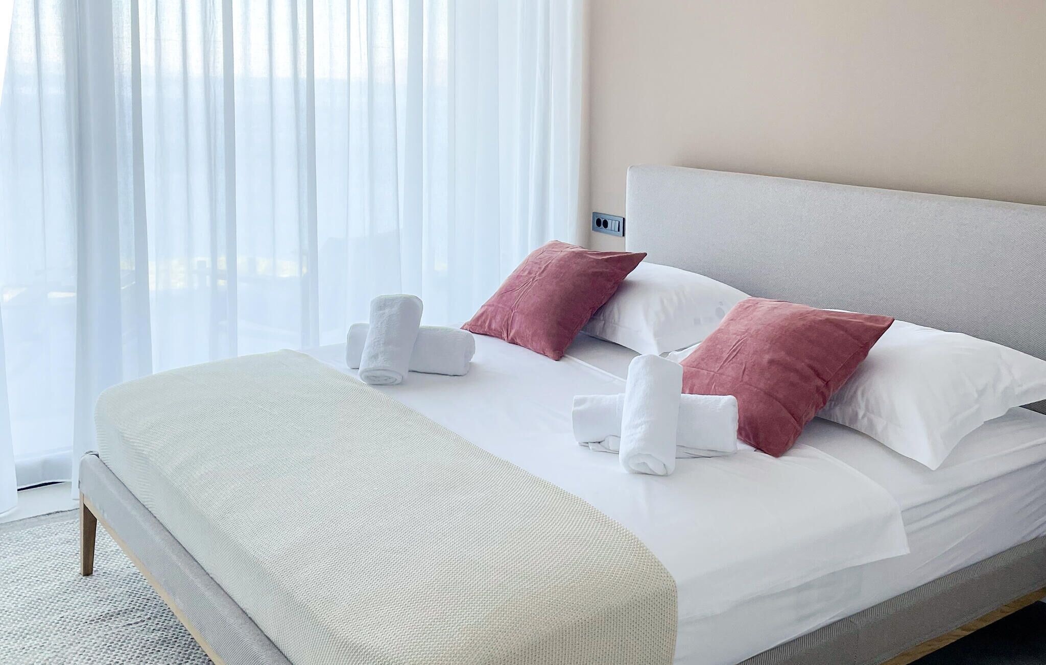 A neatly made bed with white linens and pink pillows, a bedside table with a white towel, and a window with sheer curtains.