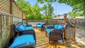 A wooden deck with wicker furniture and blue cushions, surrounded by a wooden fence and trees. - Charming 3-Bedroom House with WiFi, AC & Hot Tub in Brilliant Crystal Beach (Fort Erie)