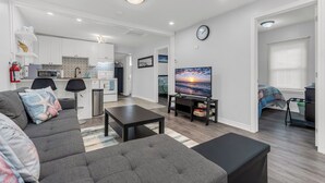 A modern living room with a sofa, coffee table, and TV. A kitchen is visible in the background with white cabinets and a tiled backsplash. - Charming 3-Bedroom House with WiFi, AC & Hot Tub in Brilliant Crystal Beach (Fort Erie)