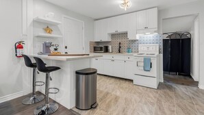 A modern kitchen with white cabinetry, a stainless steel trash bin, and a bar area with two black stools. - Charming 3-Bedroom House with WiFi, AC & Hot Tub in Brilliant Crystal Beach (Fort Erie)