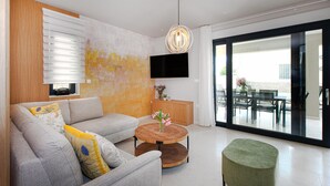 A modern living room with a grey sofa, a wooden coffee table, and a green ottoman. A flat-screen TV is mounted on the wall, and a dining area is visible through a glass door.