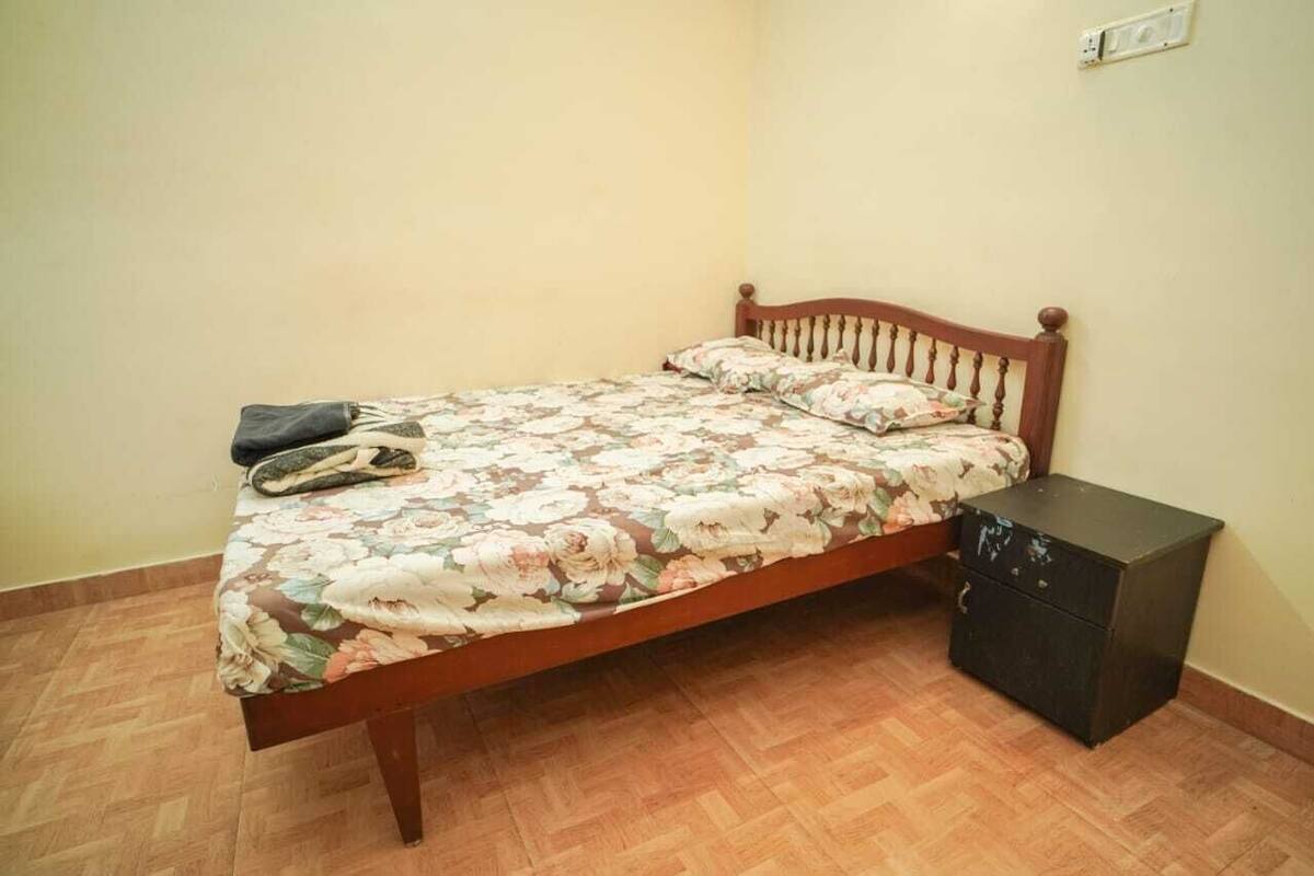 Uma cama de solteiro com colcha floral e almofadas, cabeceira de madeira e uma pequena mesa de cabeceira preta com gavetas.