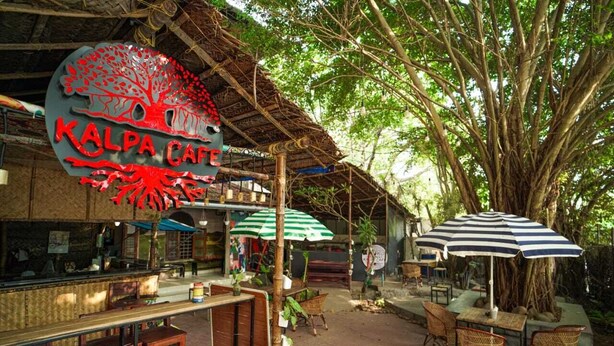 Un café al aire libre rústico con un árbol grande, mobiliario de madera y un letrero que dice "Kalpa Cafe".