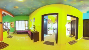 Una habitación de colores vivos con un gran pilar naranja, una pared verde y una pared amarilla. Se ve una ventana con vistas al exterior, una silla y una pequeña mesa con una planta encima.