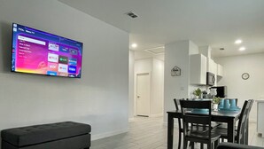 Un salon moderne doté d’un téléviseur à écran plat diffusant des services de streaming, d’un canapé gris foncé, d’un coin repas avec une table et des chaises, et d’une cuisine en arrière-plan.