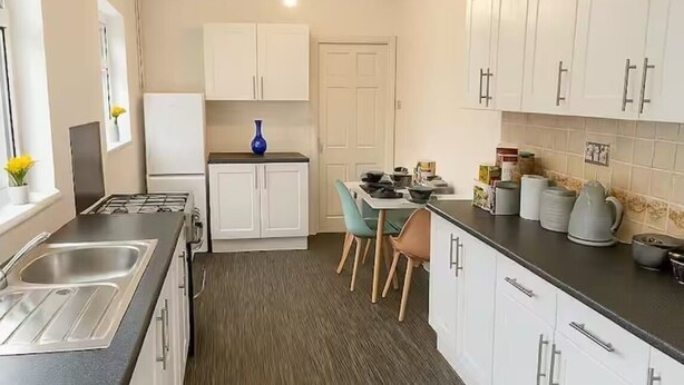 Una cocina moderna con armarios blancos, encimera negra y suelo de madera oscura. Hay un comedor con una mesa y sillas.