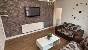 Una sala de estar moderna con pared de piedra, un televisor de pantalla plana, una araña, una mesa de centro blanca y un sofá marrón con cojines florales.