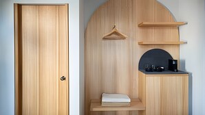 Una habitación con puerta de madera, un banco con cojín, perchero y una pequeña zona de kitchenette con cafetera.