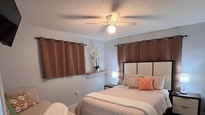 A bedroom with two beds, a ceiling fan, a TV mounted on the wall, and a small table with a lamp.