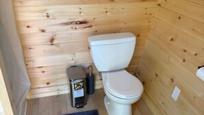 A small, wooden-paneled bathroom with a toilet, a trash bin, and a blue rug on the floor.