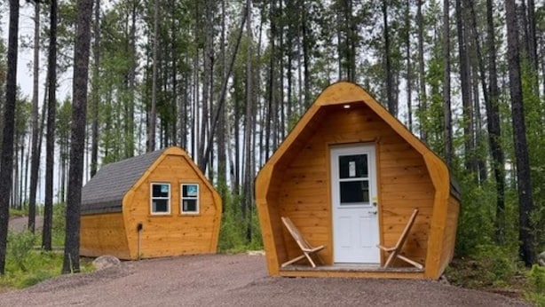 Deux cabanes en bois, avec une allée en gravier, entourées de grands arbres.
