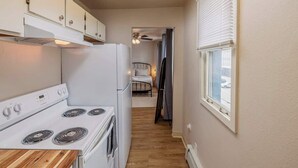 Cocina compacta con estufa, refrigerador y alacenas blancos. Una ventana con persianas está a la derecha, y una puerta da a un dormitorio con cama y mesita de noche.