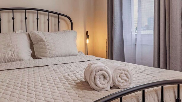 Una cama con ropa de cama bien hecha, con cubrecolcha acolchado, dos almohadas y toallas enrolladas sobre una estructura metálica, en una habitación con ventana y cortinas.