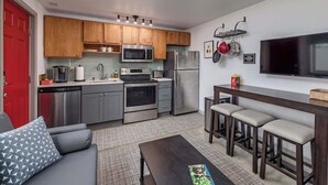Cocina compacta con electrodomésticos modernos, comedor con taburetes de bar y televisor de pantalla plana montado en la pared.