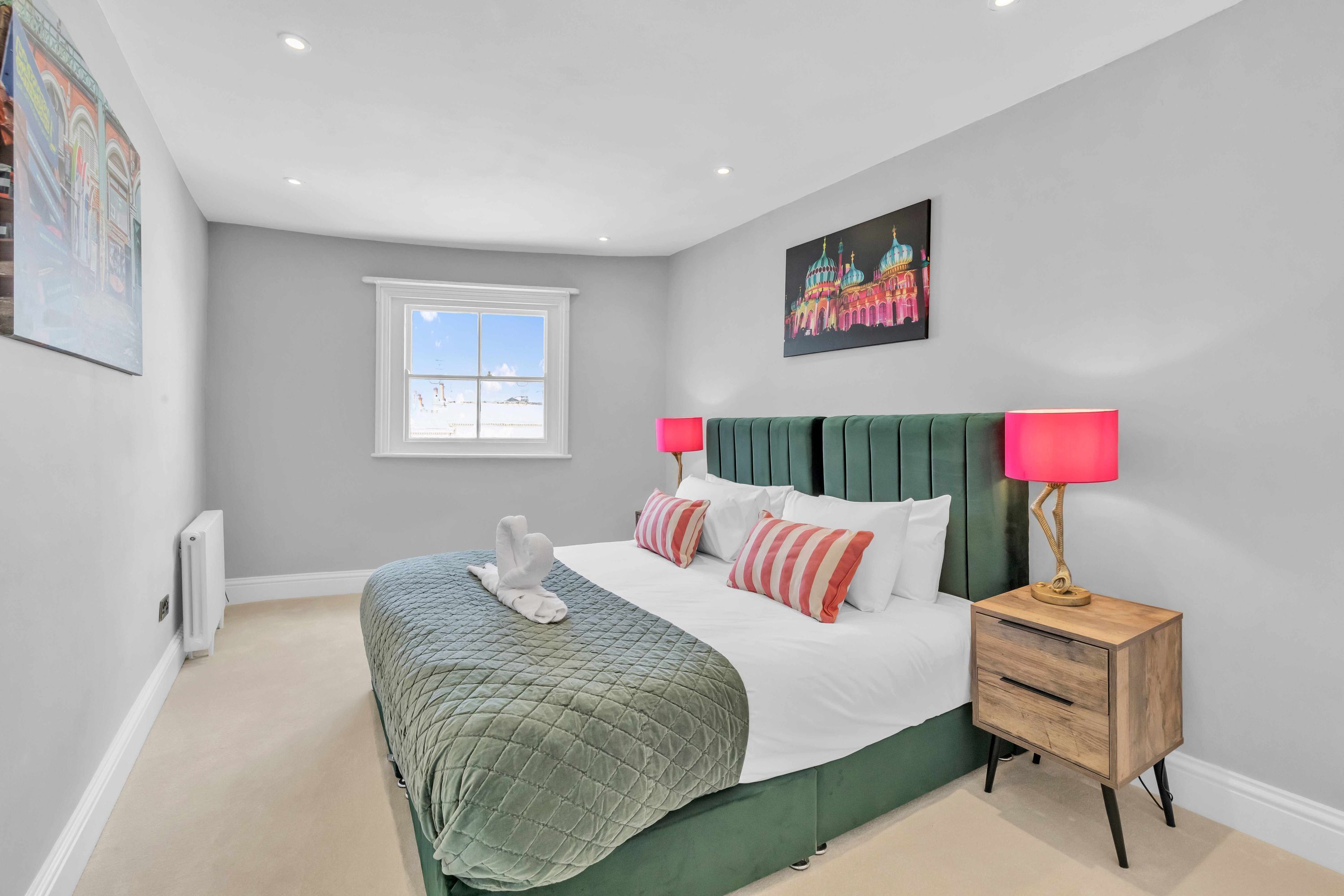 A bedroom with a green bed, white bedding, a pink lamp, and a painting of a building.