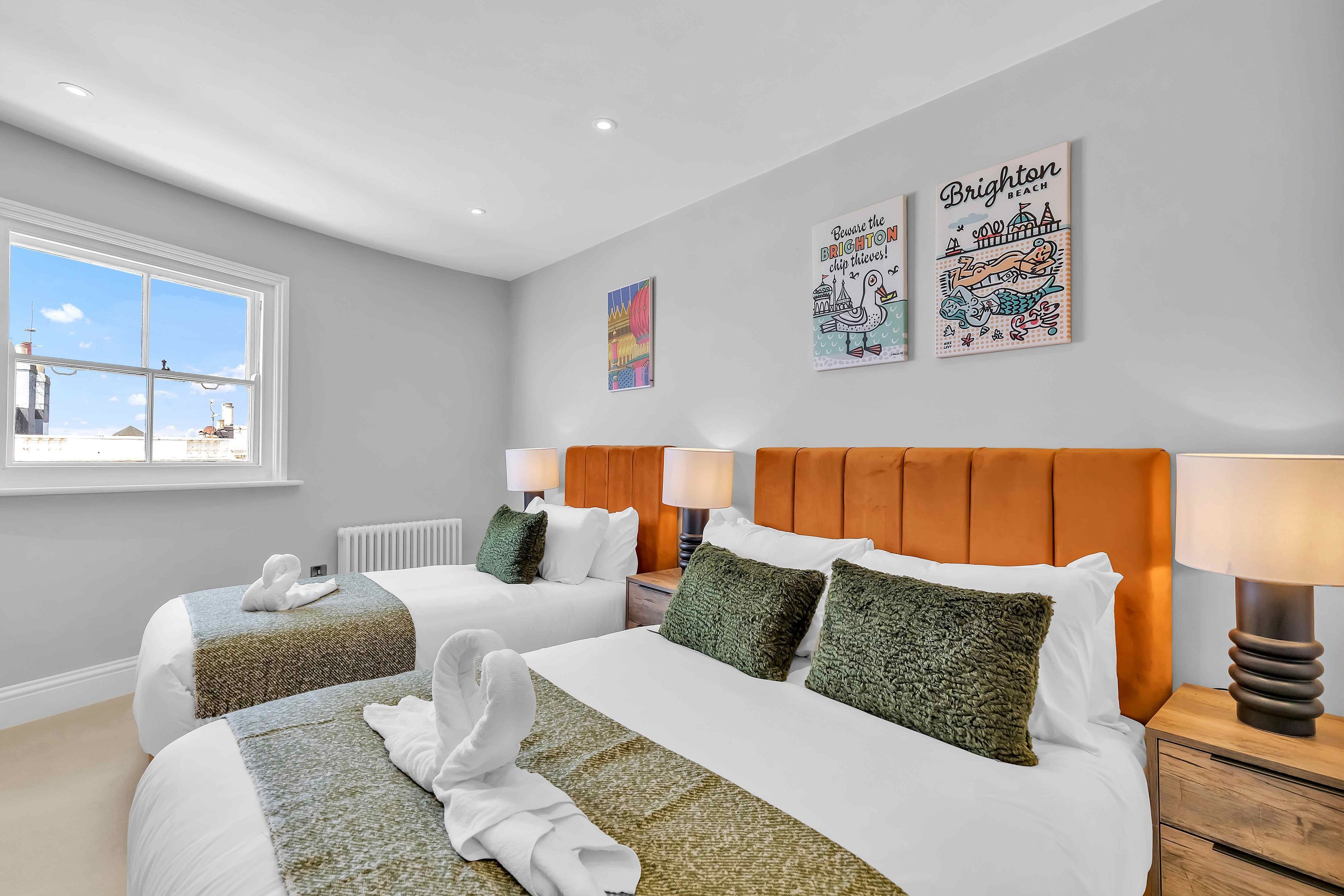 A hotel room with two beds, a window with a city view, and artwork on the wall.