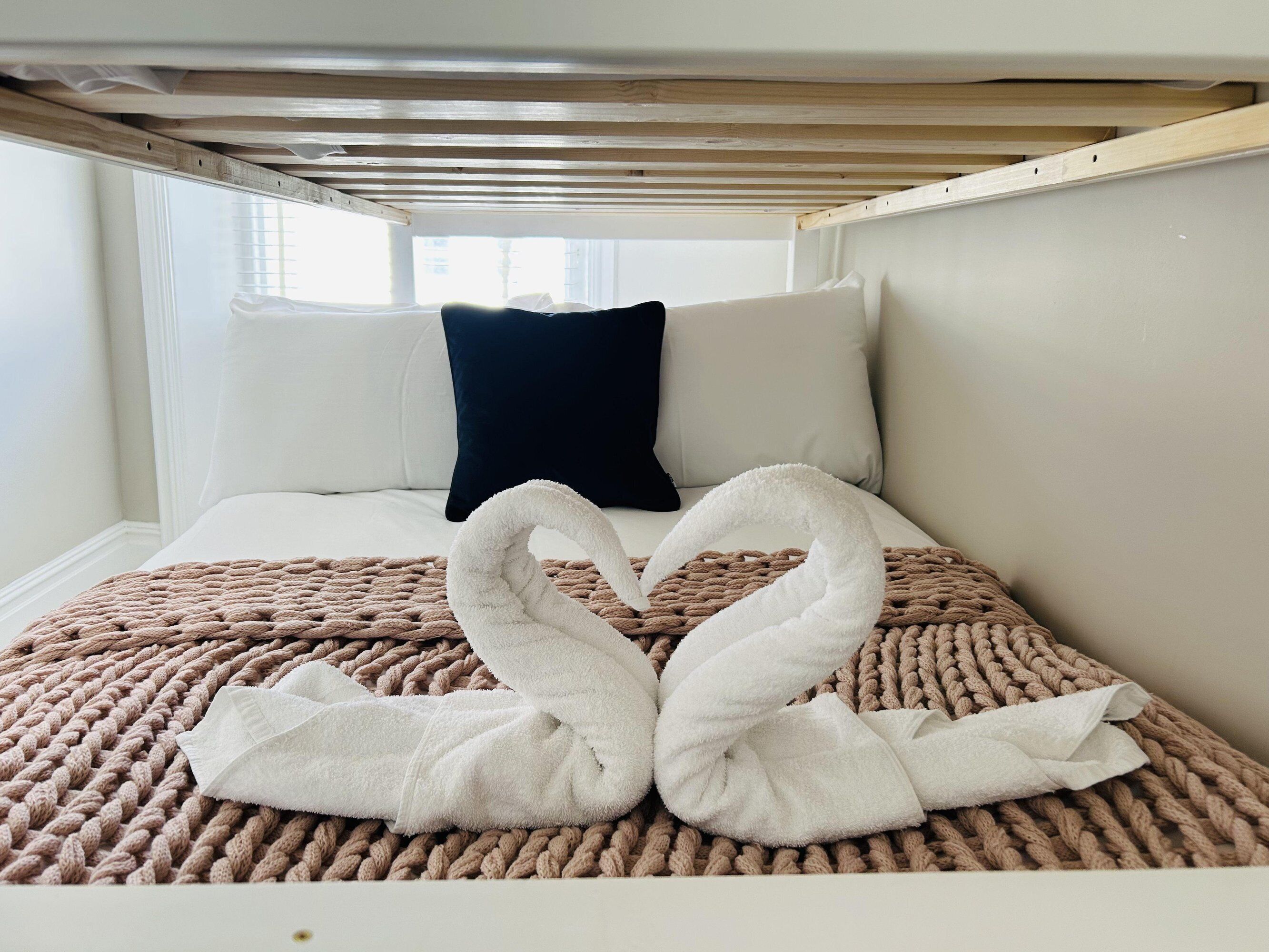 A bed with a brown knitted blanket, white pillows, and a white towel arranged in a heart shape.