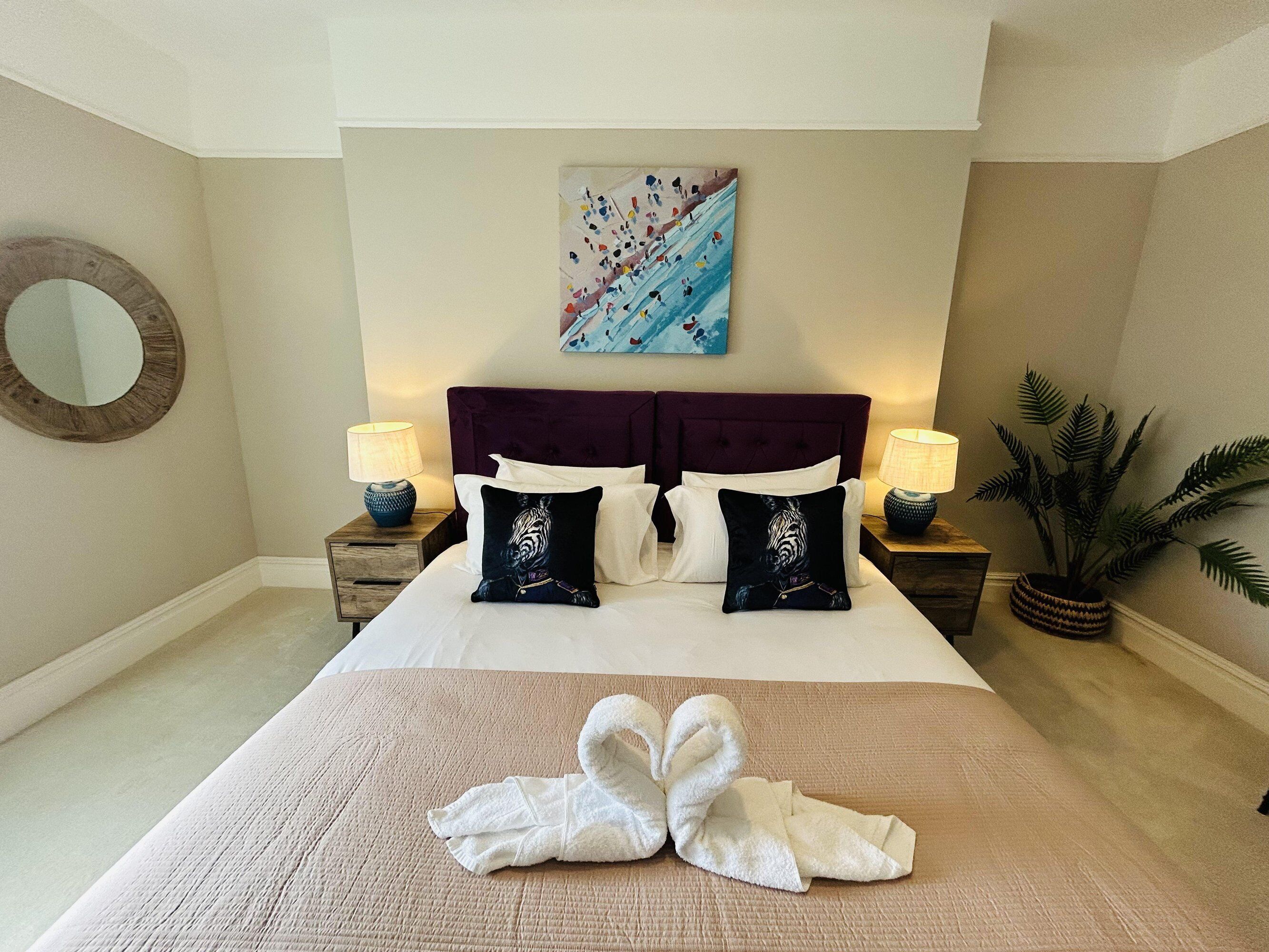 A hotel room with a large bed, two nightstands, a round mirror, a painting, and a potted plant.