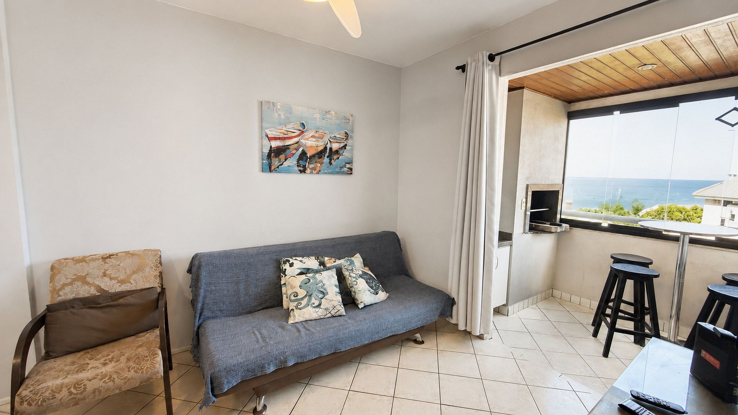 A small living area with a sofa, a chair, a painting, and a view of the sea through a window.