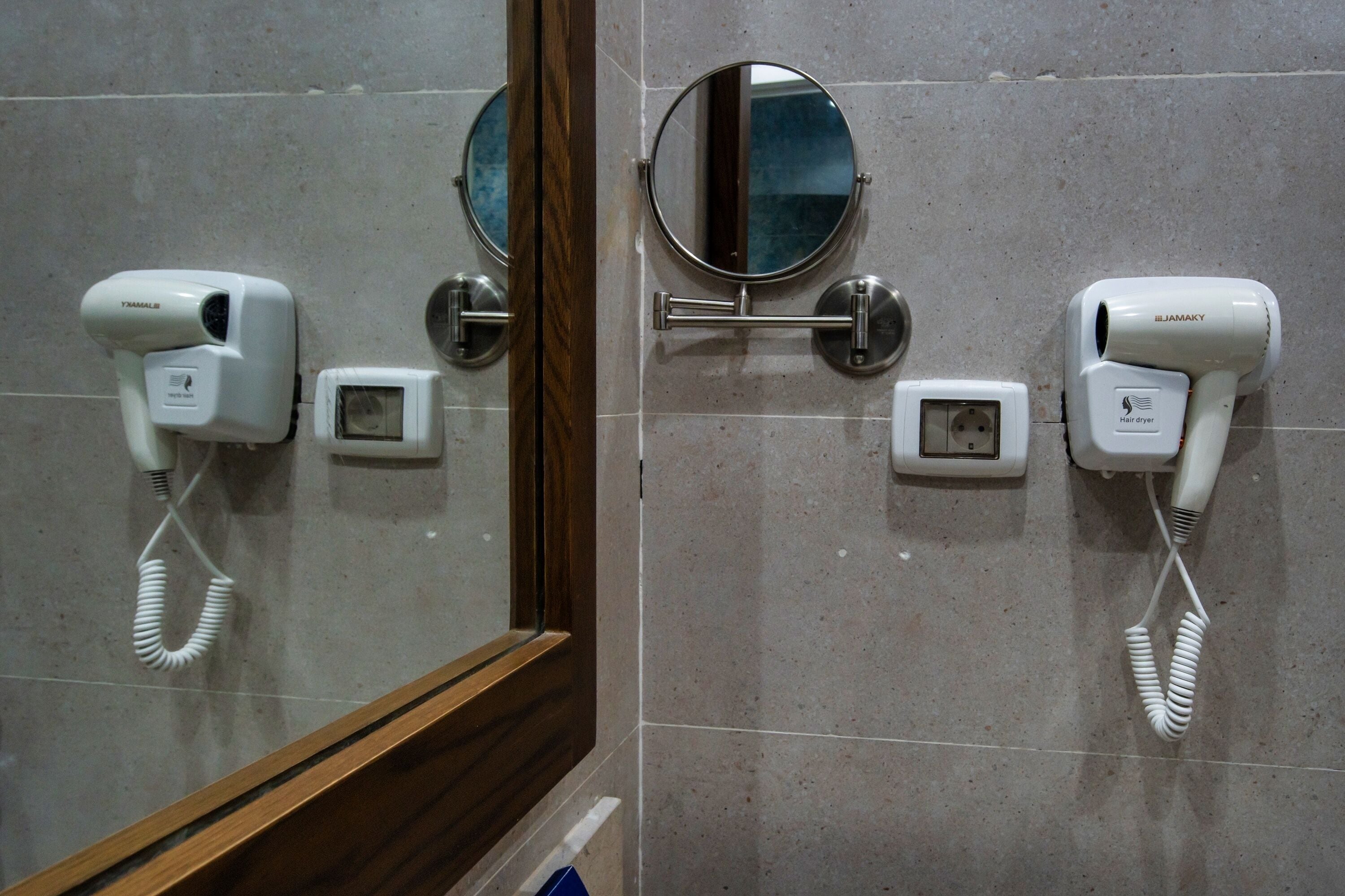 Un baño con dos secadores de pelo fijados a la pared, un espejo pequeño y un espejo con marco de madera.