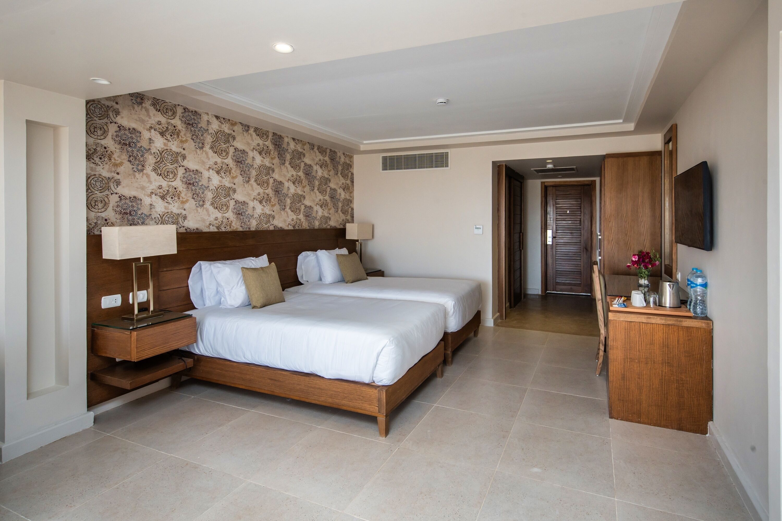 A hotel room with two beds, a wooden headboard, a nightstand, a desk, and a wall-mounted TV.