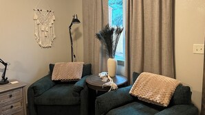 Une pièce chaleureuse avec deux fauteuils vert foncé, une petite table ronde ornée d’un vase contenant des plantes séchées et une décoration murale en macramé.