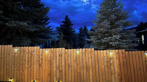 Une clôture en bois avec des lattes verticales régulièrement espacées, un décor d’arbres à feuilles persistantes et un ciel nocturne nuageux.