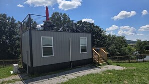 A small, single-story cabin with two windows and a covered outdoor area with a red umbrella. - Tiny Home with Rooftop Deck (Pikeville)