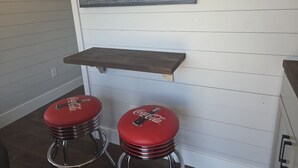 A kitchen with white paneling, two red bar stools with Coca-Cola branding, and a wooden shelf above them. - Tiny Home with Rooftop Deck (Pikeville)