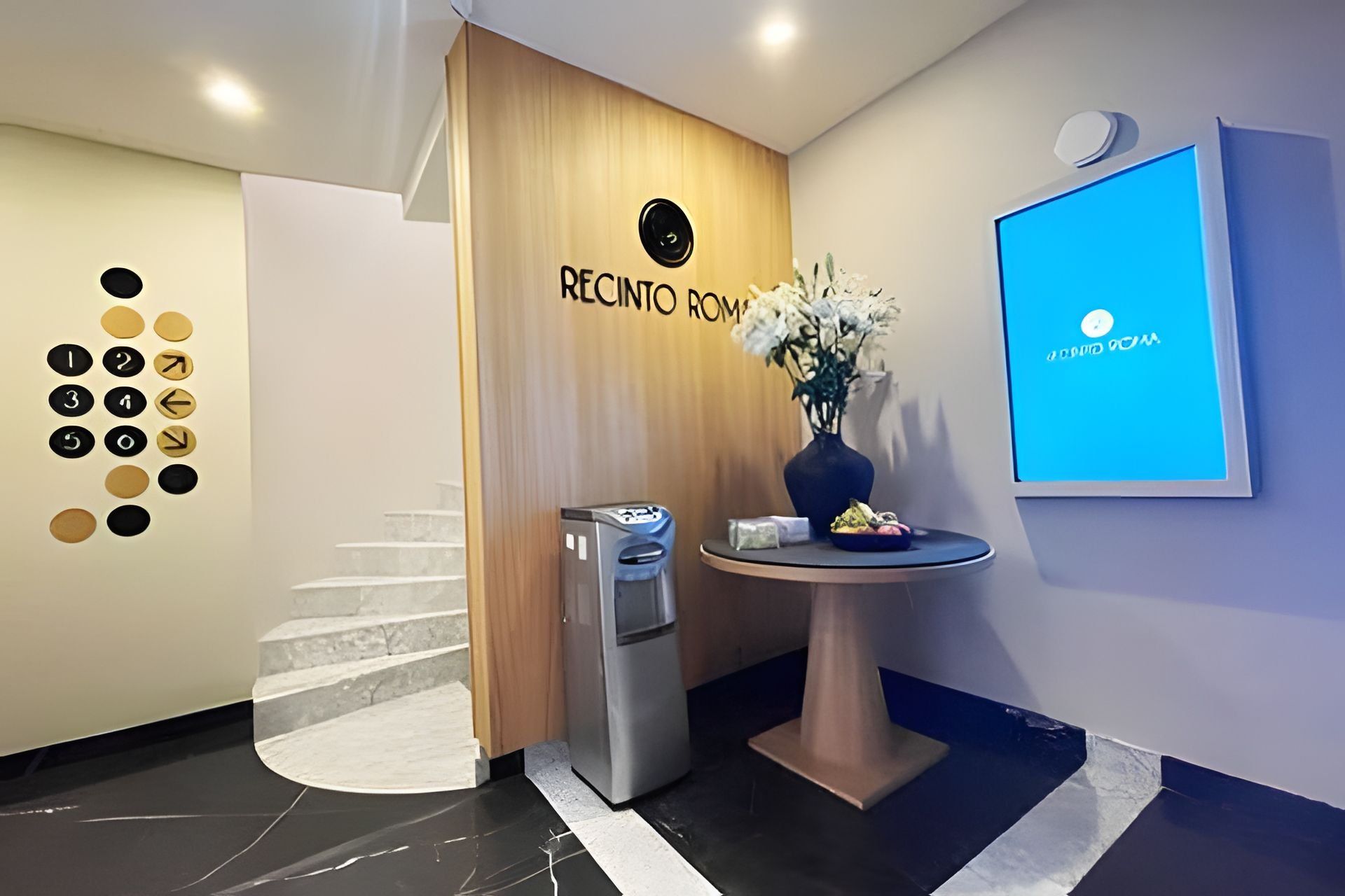 Zona de recepción con pared de paneles de madera, una pantalla digital mostrando un mensaje de bienvenida, una pequeña mesa con un jarrón de flores y un cesto de basura.
