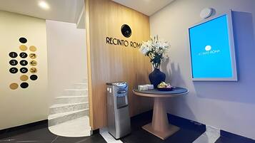 Zona de recepción con pared de paneles de madera, una pantalla digital mostrando un mensaje de bienvenida, una pequeña mesa con un jarrón de flores y un cesto de basura.
