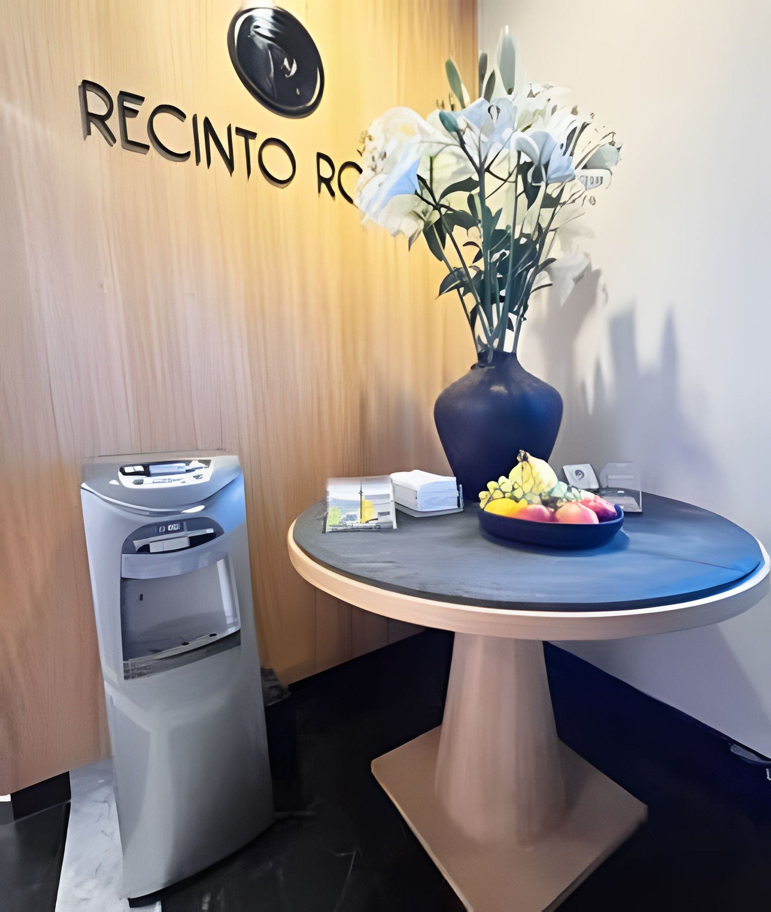 Un mostrador de recepción con un jarrón de flores, un dispensador de agua y una mesita con un frutero.