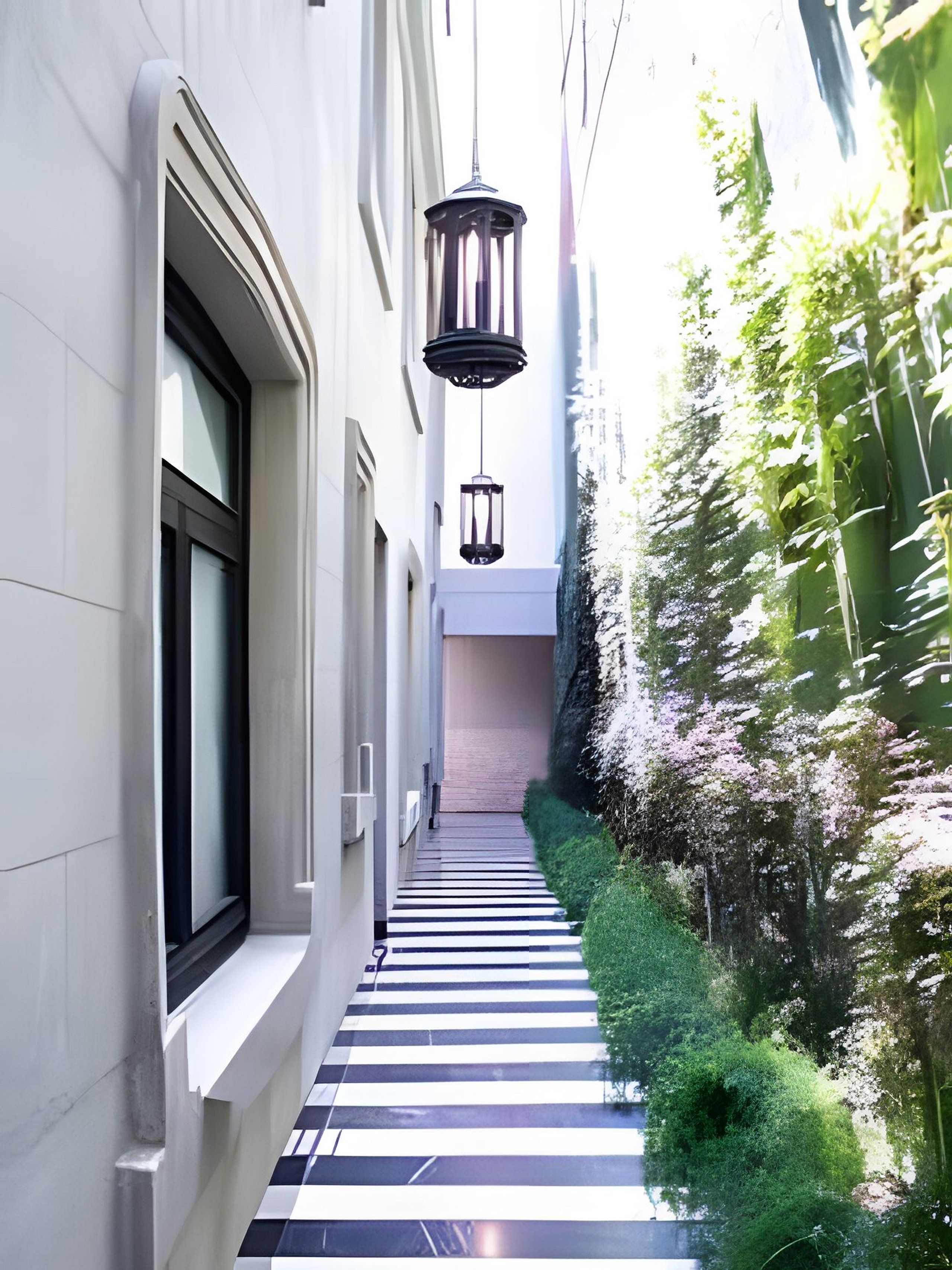 A narrow, tiled pathway flanked by buildings and greenery, leading to a door at the end.