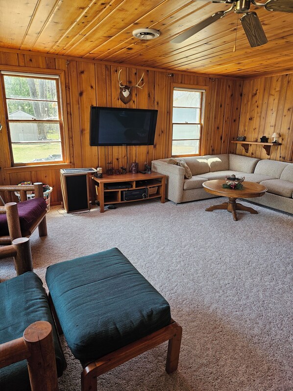 A cozy cabin interior with a wooden ceiling, a mounted TV, a sofa, a coffee table, and a fireplace.