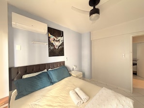 Un dormitorio con cama, ventilador de techo, equipo de aire acondicionado, una obra de arte en la pared y una puerta que da a otra habitación.