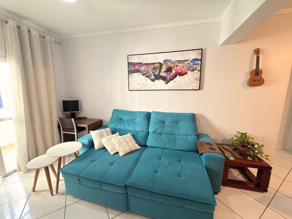 Un canapé de couleur turquoise avec des coussins, une guitare en bois, un tableau abstrait encadré, une petite table avec une plante et un téléviseur posé sur un meuble en bois.