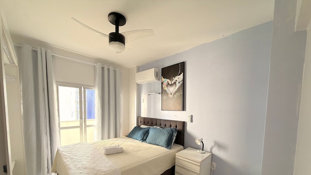 Ein Schlafzimmer mit einem Bett, einem Nachttisch, einem Deckenventilator, einem Fenster mit Vorhängen und einem an der Wand hängenden Kunstwerk.